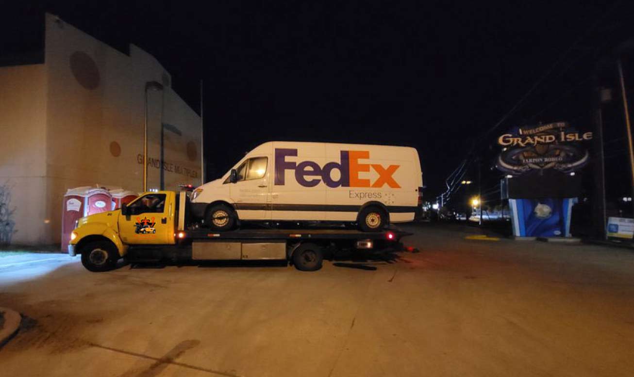 flatbed tow truck transporting a FedEx delivery van at night in a well-lit urban area.