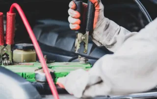 Gloved hand holding jumper cables connected to a car battery during a jump-start process.