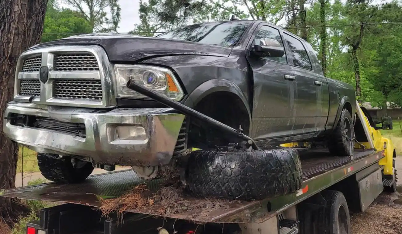 Damaged black pickup truck being towed on a flatbed truck with a loose tire and tools nearby in a wooded area.