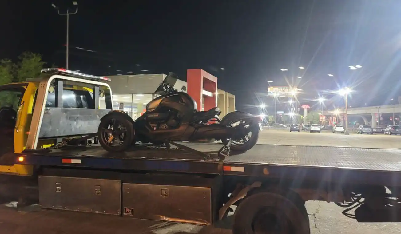 flatbed tow truck transporting a motorcycle at night in a brightly lit urban area.