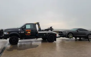 Black tow truck towing a grey sports car on a wet road under an overcast sky, providing professional towing services.