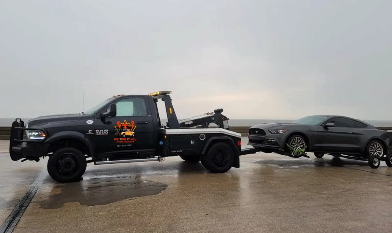 Black tow truck towing a grey sports car on a wet road under an overcast sky, providing professional towing services.