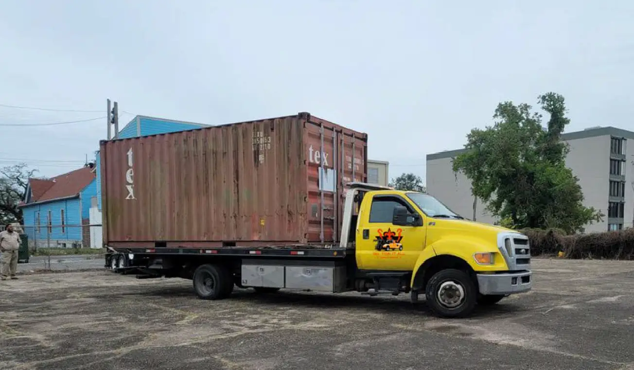 flatbed tow truck transporting a large shipping container in an urban area with residential and commercial buildings in the background.