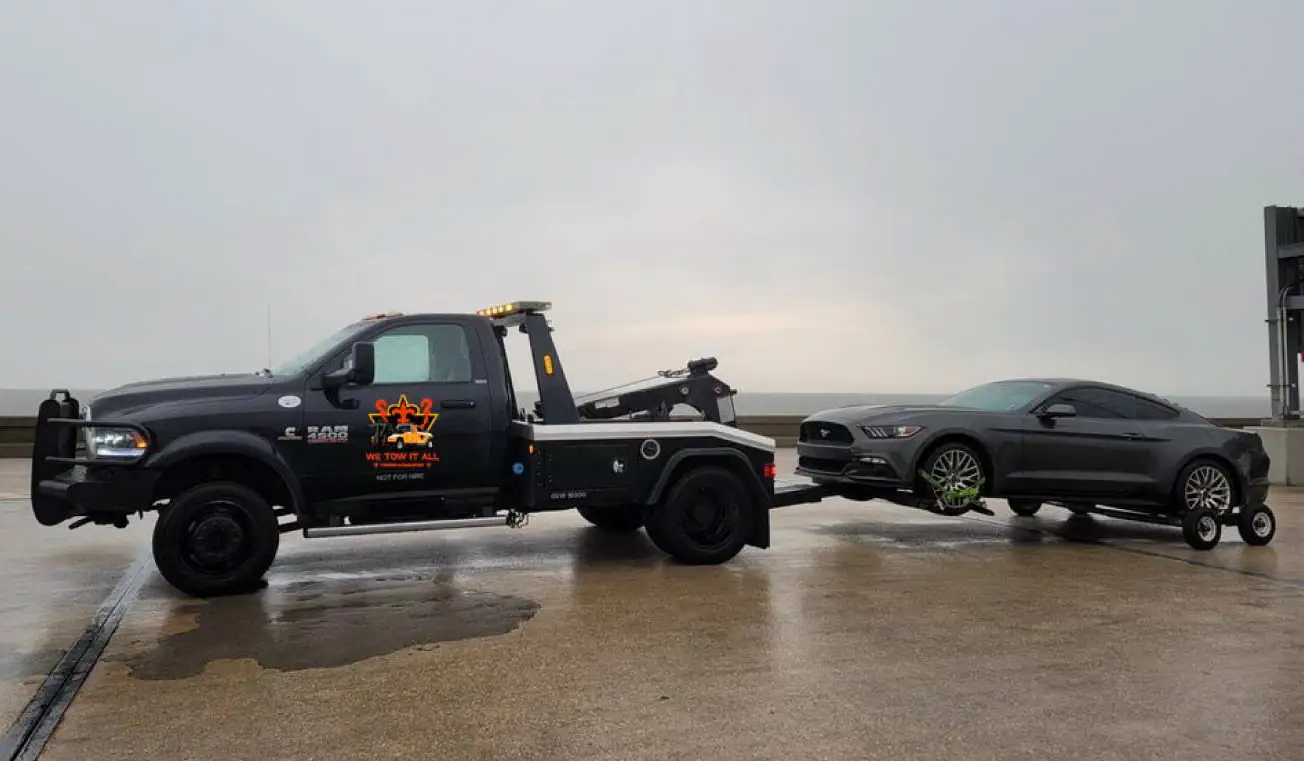 Black tow truck towing a grey sports car on a wet road under an overcast sky, providing professional towing services.