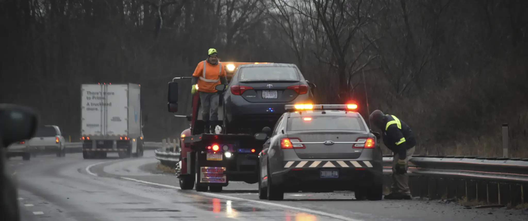 Towing service in action on a rainy highway, with a tow truck lifting a blue car while emergency response workers assist. Bright lights on the tow truck and police car illuminate the scene during the rainy weather.