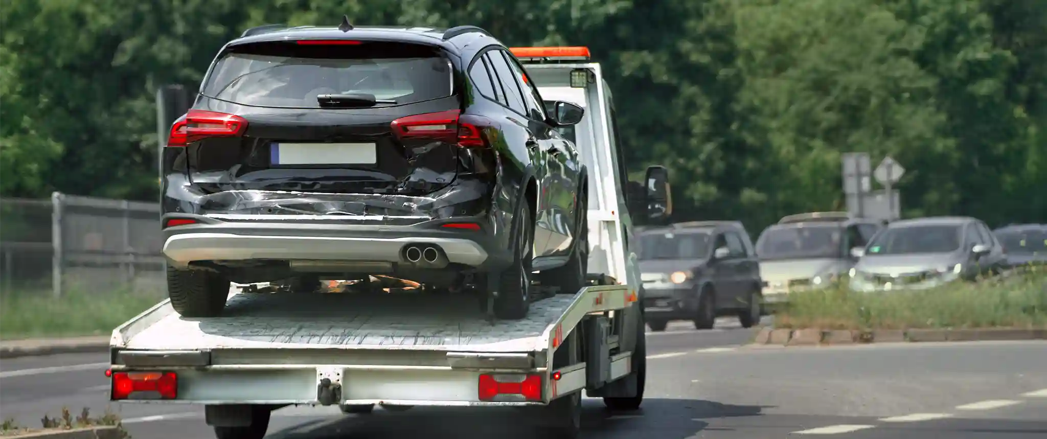 A damaged black car being towed on a flatbed tow truck along a busy road, with other cars in the background and greenery along the roadside.