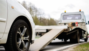 Flatbed towing truck loading a white car on the roadside with a clear sky and trees in the background.