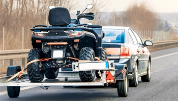 Motorcycle and ATV towing service with an ATV loaded onto a trailer behind a car, driving on a road with bare trees in the background.