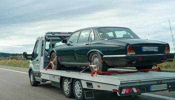 Specialty towing truck transporting a vintage green car with wheel straps on a flatbed, driving on a highway under a cloudy sky.