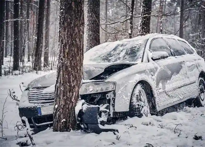 Damaged car crashed into a tree in a snowy forest, requiring accident recovery service for extraction and repair.