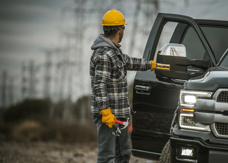 Specialty towing service with a worker in a hard hat using tools to open a truck door for towing in a remote location.