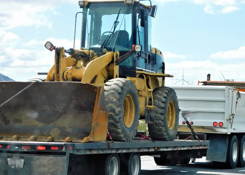 Machinery hauling service with a heavy-duty loader being transported on a flatbed trailer down the road.