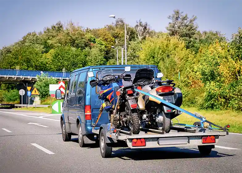 Motorcycle towing service with two bikes securely loaded on a trailer being towed by a van on a highway.