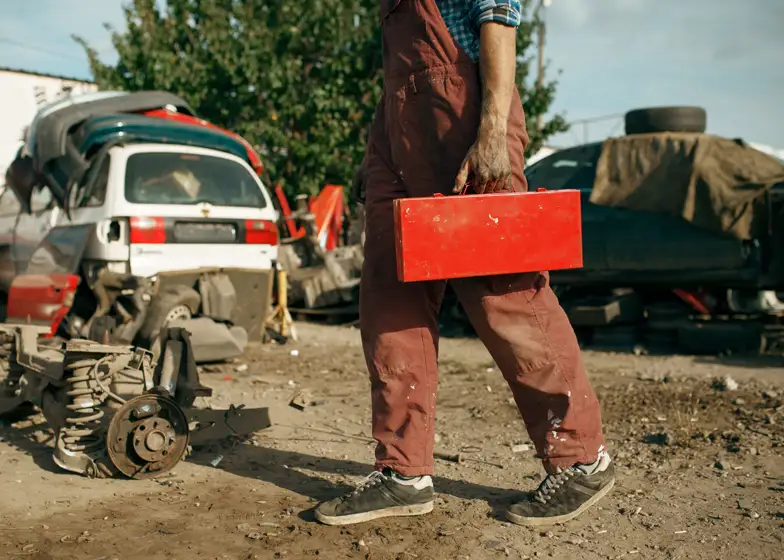 Off-road recovery service with a mechanic carrying a toolbox in a scrapyard, surrounded by vehicle parts and off-road vehicles.