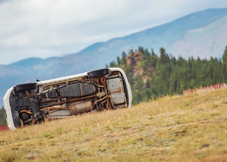 Upside-down vehicle in a grassy field, showing the undercarriage after a rollover accident, awaiting recovery in an off-road environment.